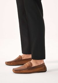 Mocasini maro din piele cu textură netedă, profil redus și detaliu decorativ de cusătură pe partea superioară. Purtați cu pantaloni negri.