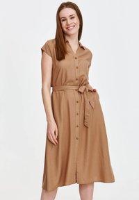 Brąsowa sukienka na krótki rękaw z guzikami, z paskiem w talii i średniej długości dołem. Gładki materiał o jednolitej fakturze i prostym kroju.