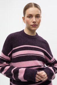 Jauna sieviete ar gaišu ādu un atpakaļ sasietiem matiem, ģērbusies purpura un rozā svītrainā pūkainā džemperī, rokas sakrustotas priekšā.