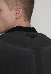 Zwarte bomberjack met een gestructureerde afwerking, voorzien van een geribde kraag en de geperste merknaam "MULTIPLY APPAREL" op de achterkant.