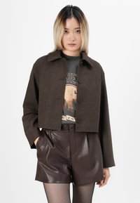 Brun kort jacka med en texturerad tyg, knäppning och spetsig krage, som bärs över en grafisk t-shirt och bruna shorts i konstläder.