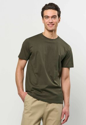 Ung mand smiler iført olivengrøn kortærmet t-shirt og beige bukser med den ene hånd i lommen, mod en ensartet lys baggrund.
