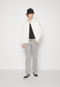 Hvit jakke over en svart t-skjorte, grå jeans og svarte sneakers med hvite såler. Modellen har på seg en svart caps og står mot en hvit bakgrunn.