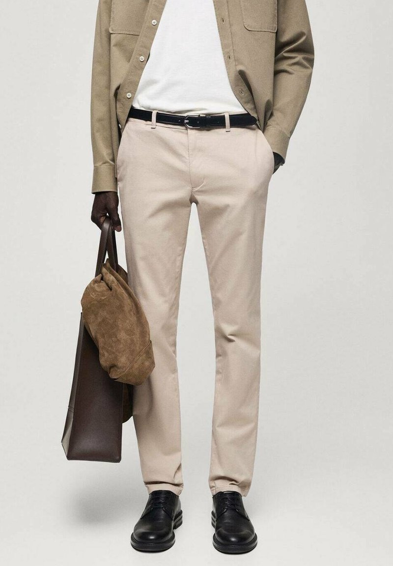 Chinos beiges à coupe droite, portés avec une chemise claire. Un sac en daim marron est tenu dans une main, assorti à des chaussures noires.