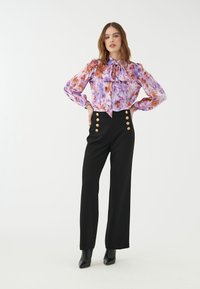 Luftige lila und orange Blumenbluse mit Volantkragen, kombiniert mit schwarzen hoch-taillierten Hosen mit goldenen Knopfakzenten und tailliertem Schnitt.