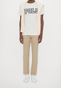 Vit T-shirt med "POLO" tryckt i marinblått, krämfärgad stickad väst, beige byxor och vita sneakers. Helhetsintrycket är avslappnat och stilrent.