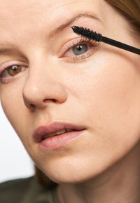 Zwarte mascaraborstel die langs lange, donkere wimpers strijkt; close-up van een lichte huid, subtiele lipkleur en een neutrale achtergrond.