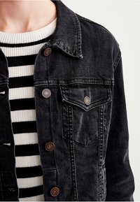 Zwarte spijkerjack met knopen aan de voorkant, twee borstzakken en een licht vervaagde textuur, gedragen over een zwart-wit gestreepte trui.