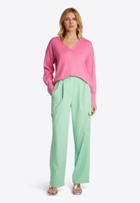 Rosa V-Ausschnitt Pullover kombiniert mit mintgrünen, weiten Hosen mit seitlichen Taschen. Das Outfit wird durch silberne Riemchensandalen ergänzt.