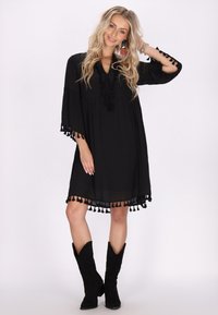Schwarzes Boho-Style Kleid mit weit ausgestellten Ärmeln und Quastenakzenten am Saum. Hergestellt aus leichtem Stoff, mit lockerer Passform und V-Ausschnitt.