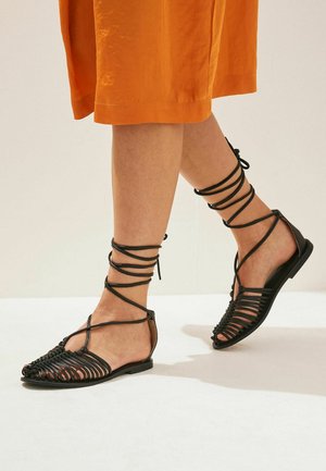 Pieds portant des sandales plates noires à brides avec des liens enveloppant la cheville, associés à une jupe orange mi-longue sur un fond neutre.