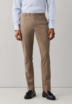 Hombre que lleva pantalones ajustados marrón claro, camisa azul clara a rayas y mocasines negros con borlas, de pie frente a un fondo liso.