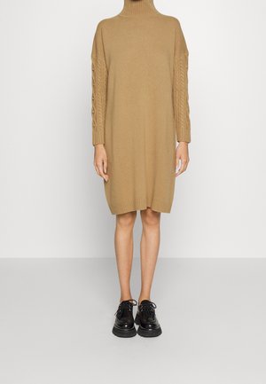 Persoon draagt een camelkleurige coltrui-jurk met kabelgebreide mouwen en zwarte stevige veterlaarzen, staand op een grijze vloer.