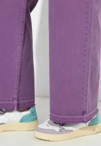 Jeans de pierna ancha morados con dobladillos deshilachados, combinados con zapatillas multicolores que presentan acentos en blanco, púrpura claro y verde azulado.