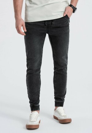 Zwarte tapered jeans met elastische manchetten, denimstructuur, voorzakken en een slim fit, gecombineerd met witte en tan sneakers.