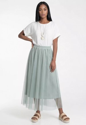 Kobieta z długimi czarnymi włosami ubrana w biały top z krótkim rękawem, długą jasnobieżą spódnicę tiulową, beżowe sandały i długi naszyjnik z koralików na białym tle.