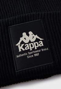 Svart ribbestrikket lue med et vevd merke som har hvit tekst "Kappa" og to sittende figurer, sammen med "Authentic Sportswear Brand" og "Since 1967."