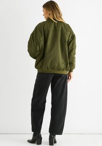 Olive green bomber jacket with a relaxed fit, gathered detailing on sleeves, and ribbed cuffs. Paired with high-waisted black denim pants.
