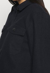 Merinopaita, tummansininen, jossa on napillinen kaulus, kaksi rintataskua ja etunapit. Pehmeä rakenne ja yksivärinen muotoilu.