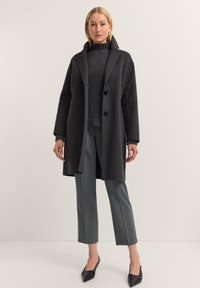 Schwarzer Wollmantel mit großen Revers, doppelten Knöpfen und Seitentaschen. Über einem dunklen strukturierten Strickpullover und hellgrauen Hosen getragen.