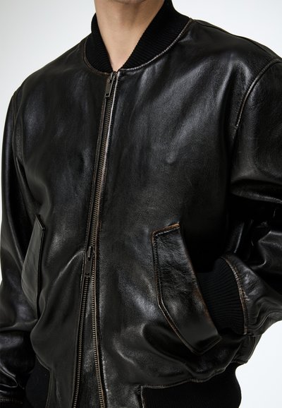 Férfi fekete bőr bomberdzsekiben, bordázott gallérral, mandzsettákkal és két ferde első zsebbel, egyik kezét a zsebében tartva áll.