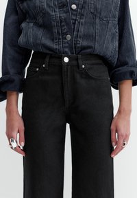 Osoba nosząca czarne jeansy z wysokim stanem i podwiniętą, ciemną koszulę dżinsową, z widocznymi srebrnymi pierścieniami na obu dłoniach po bokach.