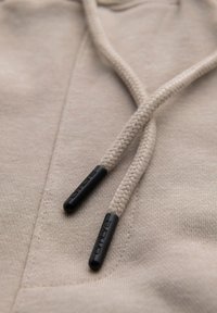 Gros plan sur le tissu beige d'un sweat à capuche avec des cordons tressés beiges se terminant par des embouts noirs portant le texte "Ombre".