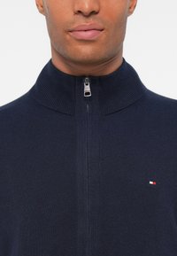 Marineblå hettegenser med høy krage, laget av ribbestrikket materiale, med en liten logo nederst til venstre. Glatt tekstur, tettsittende design.