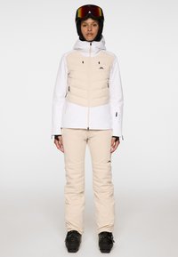 Strój narciarski z białą kurtką z beżowymi pikowanymi panelami, w połączeniu z beżowymi spodniami. Zawiera czarne akcenty oraz dopasowany kask z daszkiem.