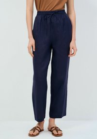 Marinblå linnebyxor med vida ben, elastisk midja och dragsko. Bärs med en brun t-shirt och bruna sandaler, vilket ger en avslappnad look.