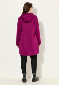Cappotto fucsia con cappuccio, dal taglio dritto, maniche lunghe e tasche laterali, abbinato a jeans neri con risvolti e stivali neri.