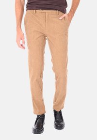 Uomo che indossa pantaloni in velluto a coste beige slim-fit con tasche laterali, abbinati a scarpe nere in pelle con lacci e una camicia marrone scuro.