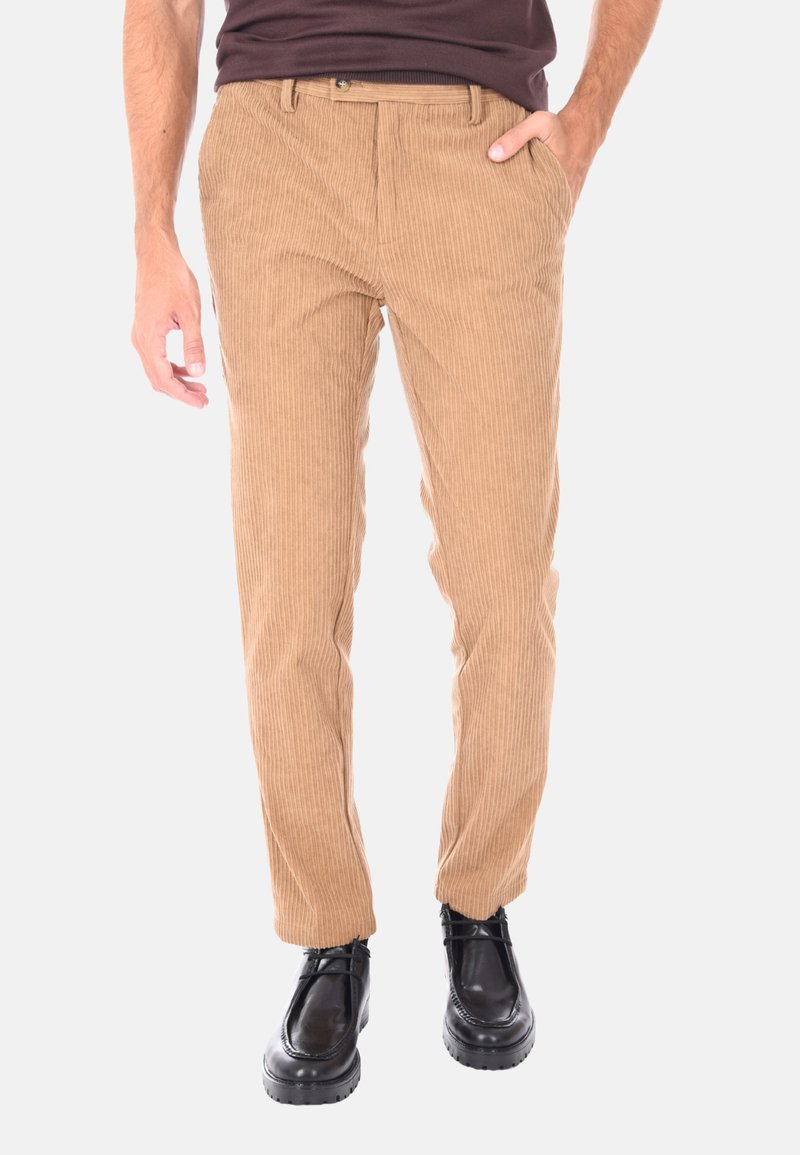 Uomo che indossa pantaloni in velluto a coste beige slim-fit con tasche laterali, abbinati a scarpe nere in pelle con lacci e una camicia marrone scuro.