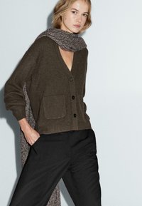 Bruine gebreide cardigan met een diepe V-hals, voorzien van knopen en twee voorkruiden, gecombineerd met een gestructureerde grijze sjaal en zwarte broeken.