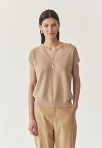 Vrouw met steil, donker haar draagt een beige trui met korte mouwen en een V-hals, bijpassende broek, gouden creolen en een ketting tegen een witte achtergrond.