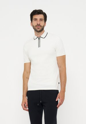 Homme aux cheveux foncés et à la barbe, portant un polo blanc à manches courtes avec un bord noir et un pantalon noir, debout devant un fond blanc uni.
