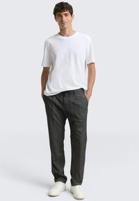 Mann steht mit den Händen in den Taschen, trägt ein weißes kurzärmeliges Hemd, dunkelgraue Hosen mit Kordelzug und weiße Turnschuhe vor einem einfachen Hintergrund.