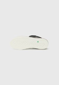 Black shoe sole with a textured pattern, featuring a white rubber base and a green accent point. Velcro strap for closure.