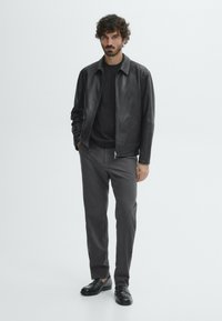 Veste en cuir noire sur un pull noir, associée à un pantalon gris et des mocassins noirs. Le modèle a des cheveux bouclés et se tient devant un fond blanc.