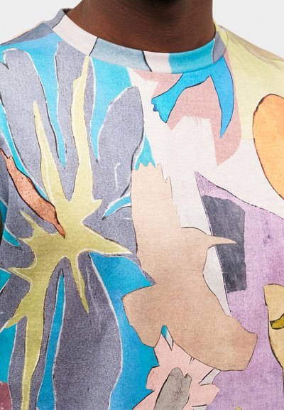 Κοντινή λήψη πολύχρωμου πουκάμισου με αφηρημένα σχήματα, συμπεριλαμβανομένης μίας μπεζ σιλουέτας πουλιού και διαφόρων μπλε, κίτρινων και μωβ σχεδίων.