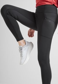 Black athletic leggings with a smooth texture and subtle stitching details, featuring a zip pocket and logo on the side, paired with gray sneakers.