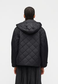 Άτομο με πλεξούδες που φοράει μαύρο πλεκτό μπουφάν με κουκούλα και σκουρόχρωμη γκρι φούστα, απεικονίζεται από πίσω με φόντο ένα απλό φόντο.