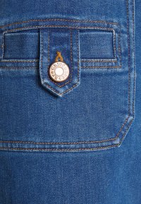 Primer plano de un bolsillo trasero de mezclilla azul con costuras naranjas detalladas y un botón de metal grabado con "Chloé See By" en la solapa.