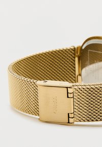 Arany színű hálós óra szíj fényes csattal, amelyen a "GUESS" márkanevének gravírozása látható. A teljes felülete sima textúrájú és fémből készült.