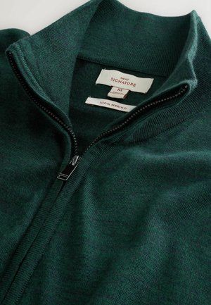 Mörkgrön merinoull zip-up-tröja med hög krage, delvis uppdraget, som visar storleks- och tygetiketter inuti kragen.