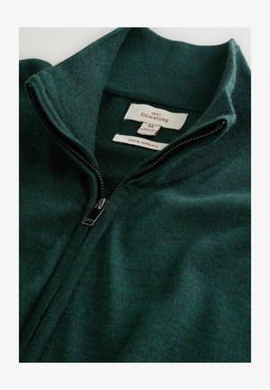 Dark green merino wool zip-up sweater with high collar, partially unzipped, showing size and fabric labels inside the collar.
