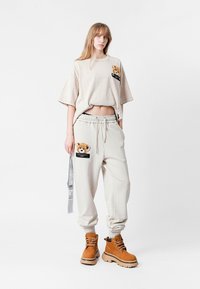 Camiseta oversized beige con un parche de oso de peluche; pantalones de chándal gris claro con detalle de oso de peluche y bolsillos laterales; botas marrones con suela gruesa.