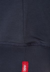 Tela de color azul marino oscuro con un acabado texturizado; presenta costuras ajustadas y una etiqueta de tela roja con la inscripción "edc" en el borde inferior.