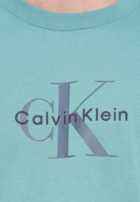 Šviesiai mėlynas medvilninis marškinėlis su ryškiu violetiniu siuvinėtu „CalvinKlein“ logotipu ir stilizuotu „CK“ akcentu.