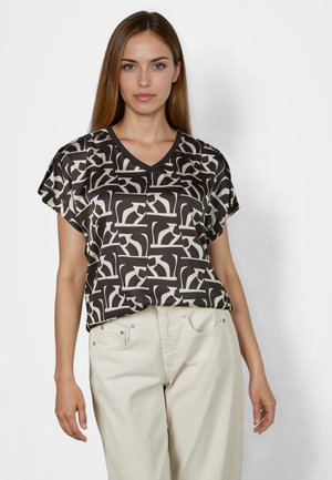 Woman wearing a short-sleeve black and white abstract patterned blouse and light beige high-waist pants, standing against a plain background.